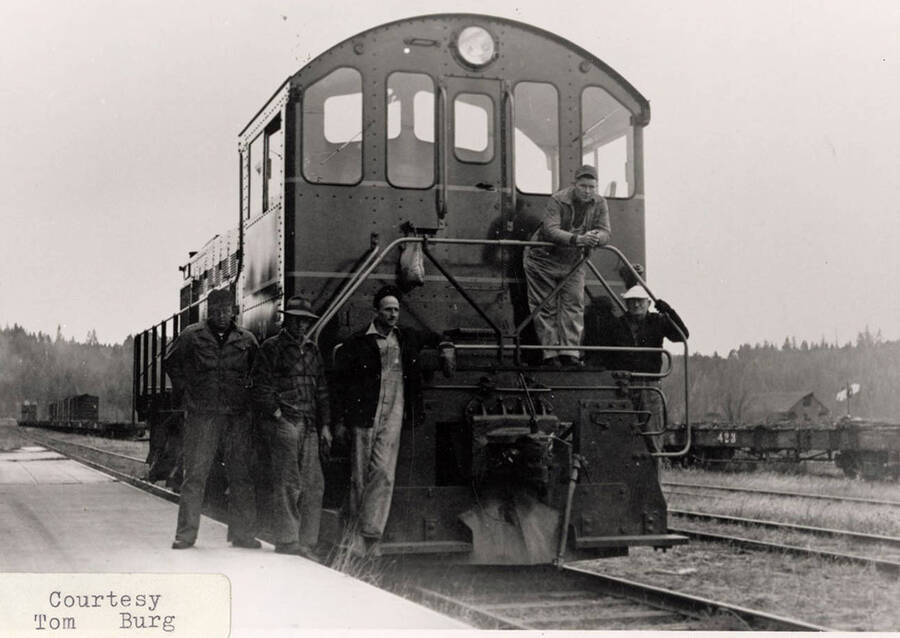 Five men standing on and next to a WI&M locomotive. Railroad cars carrying stacks of logs can be seen on other railroad tracks behind the locomotive.