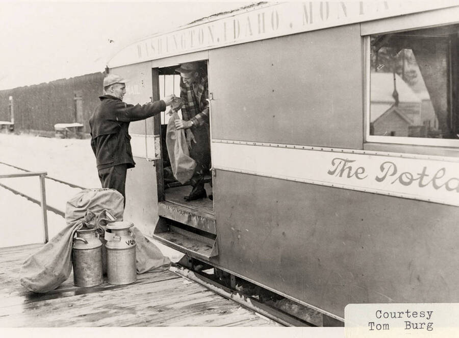 A man handing another man, who is standing inside the railroad car, a bag. The men are loading 'The Potlatcher', a streamlined passenger, mail, and express car.