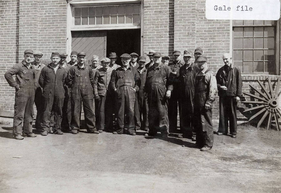 Group photo of the shop crew standing outside the entryway of a building. All the men are wearing overalls and hats.
