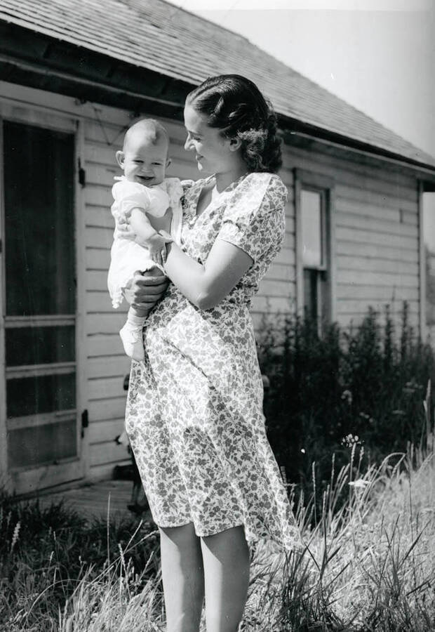 Cleora Nirk stands outside a house holding a child in her arms.