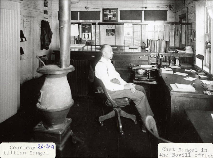 Chet Yangel sitting at a desk in the Bovill office. He is sitting next to a counter, which has books and typewriter sitting on it.
