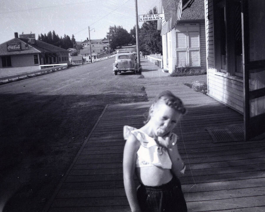 Girl stands on pine street. The confectionary can be seen on the left hand side of the street and the public library can be seen on the right. The public library was located in the old theater before moving to its current location