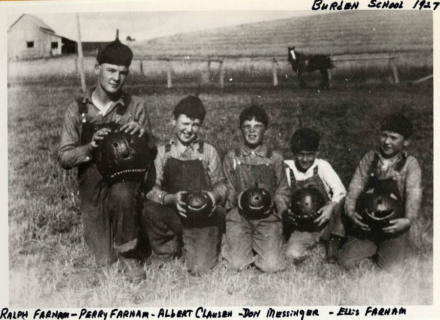 Ralph Farnam, Perry Farnam, Albert Clausen, Don Messinger, and Ellis Farnam sitting in a field near the Burden School.  Each child is holding a Jack-O-Lantern in front of them. Photograph taken in 1927.