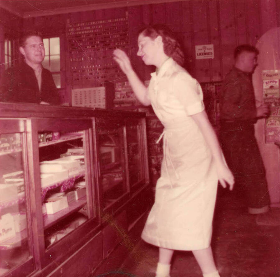 Interior view of store. Caption on back reads ''Virginia Anna Cooper working at Potlatch Confectionery. The day before I took a week off to get married. Peal Cone sneaked up to take may picture. I was trying to get away from her.'' Virginia Soncarty worked there 1955 to 1957. Located where Harvest Foods is in 2017. Small diner with soda bar, dining area, and bar in the back. Closed in mid-1970s. Not owned by the Potlatch Company, which was unusual.