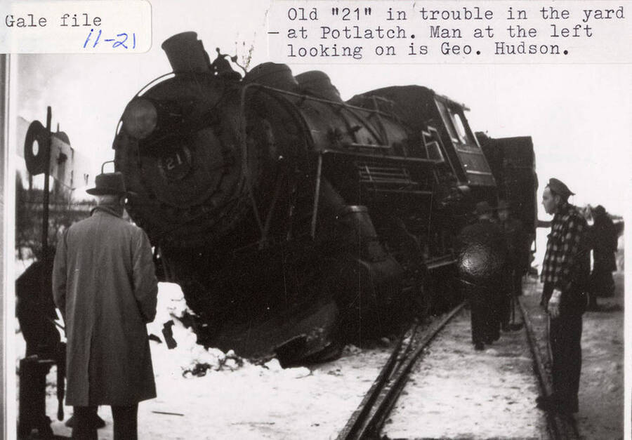 An old '21' in trouble in the yard at Potlatch, The locomotive can be seen derailed into the snow. A few men can be seen standing on the tracks and next to the locomotive. The men standing at the lest looking on is Geo. Hudson.