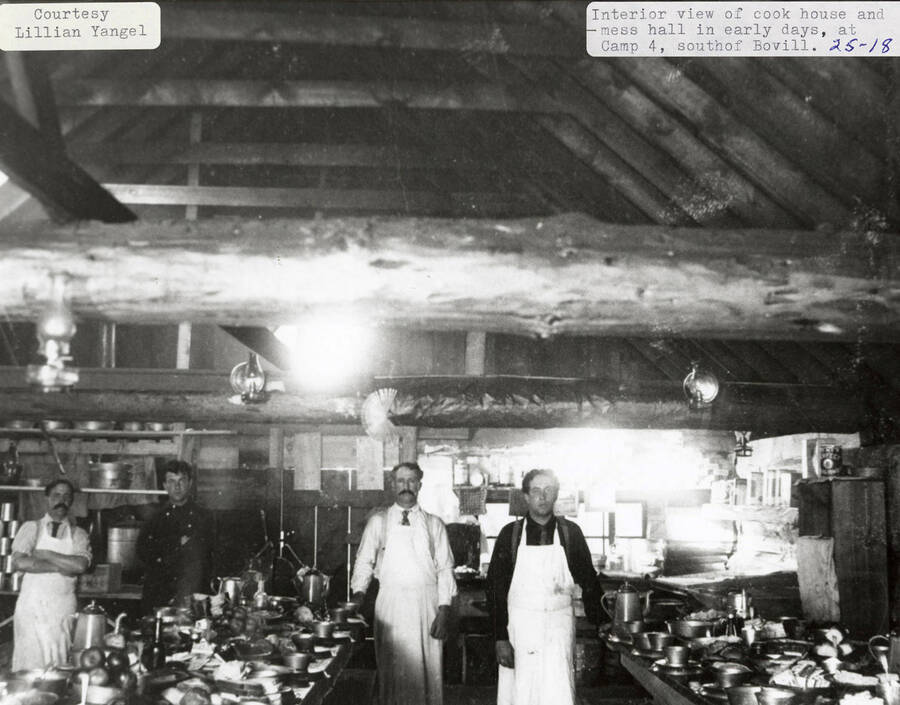 Interior view of the cook house and mess hall and Camp 4, which is located south of Bovill. Four men wearing aprons are standing next to tables stacked withy dishes.