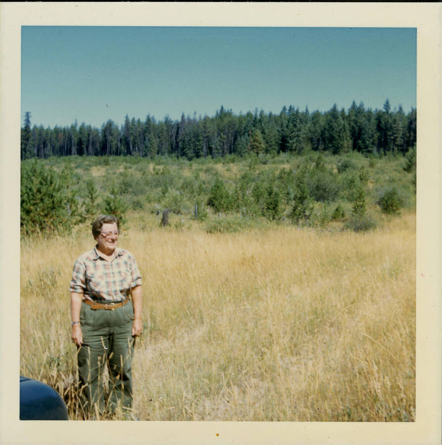 Gen Williams in plaid shirt standing in a field backed by pine trees.