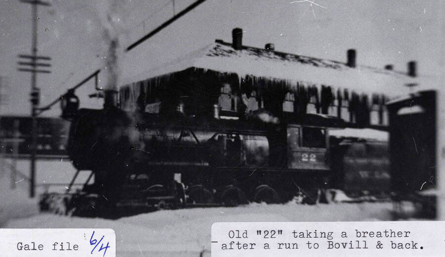 An old '22' locomotive taking a break at a station, after a run to Bovill and back. Snow can be seen surrounding the tracks and icicles can be seen hanging off the station's roof.