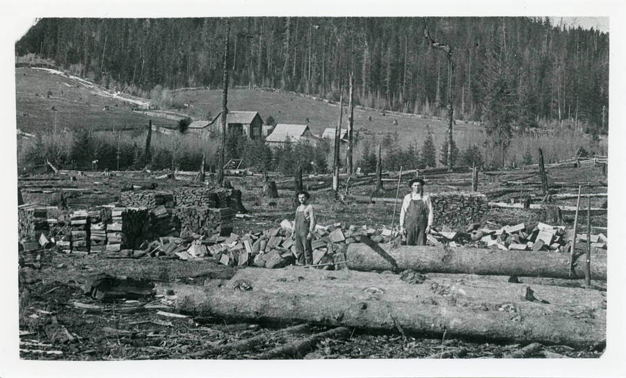 Fritz and August Lesitner with the Elmore place in the background. They're bucking logs and splitting the rounds into firewood.