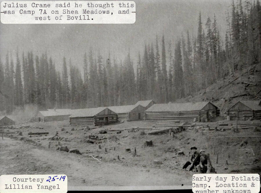 View of a Potlatch camp during the early days. The camp is thought to be Camp 7A on Shea Meadows, which is west of Bovill. Two men can be seen sitting on the camp surrounded by log cabins and tree stumps.