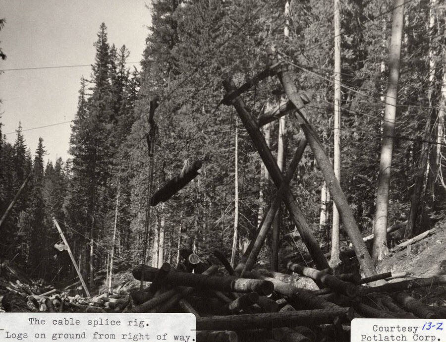 View of the cable splice rig machine. Under the machine, logs lay on the ground in stacks.