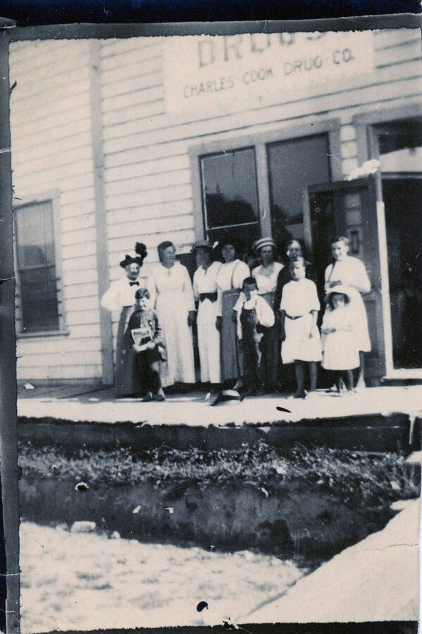 A group of unidentified people, possibly the Cook family, stands on the boardwalk in front of a drug store in Princeton.