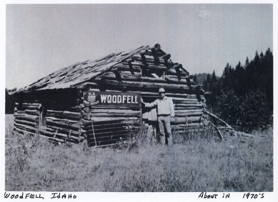 Woodfell, Idaho in the 1970's. A man stands in front of a log cabin designated as the post office. 
