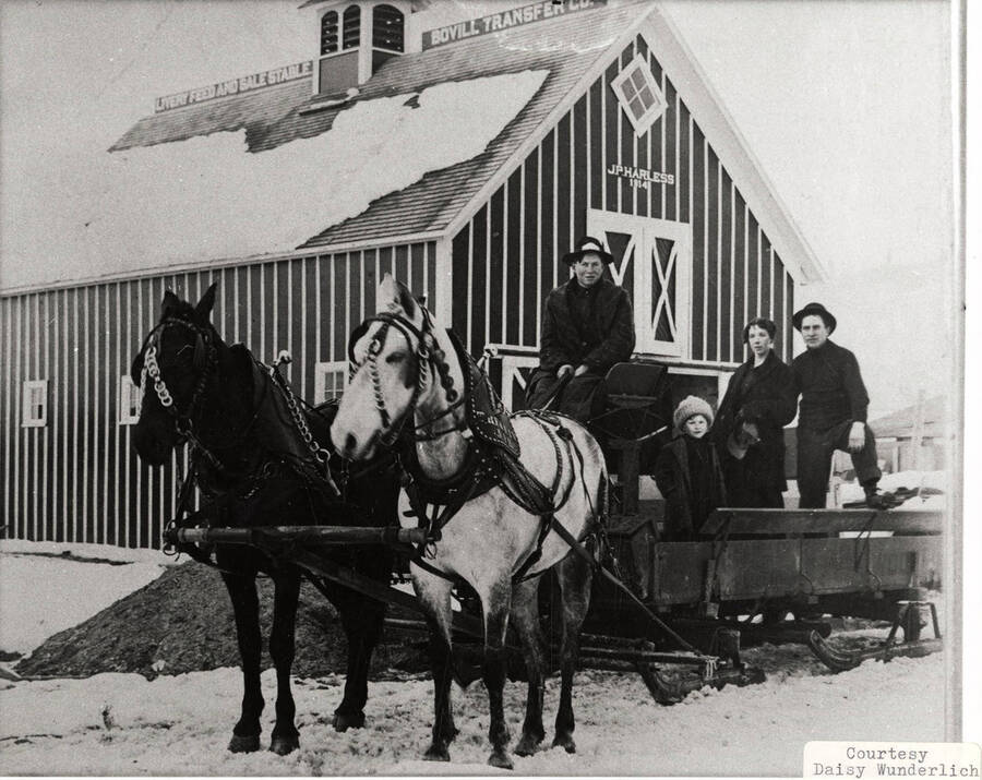 Two horses hooked up to a sleigh with four people in it. A man can be seen sitting on a high sit holding the reigns. The sleigh is barked outside of a barn, which says 'J. P. Harless' on the front.
