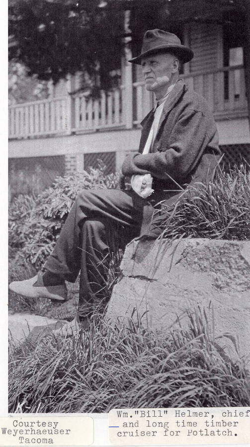 Photo of William 'Bill' Helmer, the chief and long time timber cruiser for Potlatch. He is seated on next to some plants outside of a building.