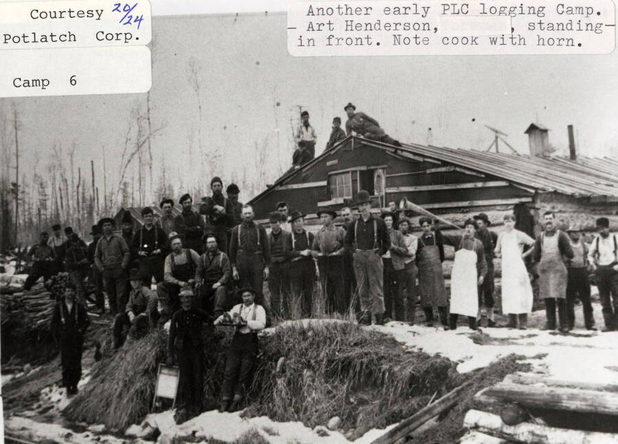 View of an early PLC logging camp. A group of people, including Art Henderson, are standing in front of a log cabin at the camp. One of the cooks can be seen holding a horn.
