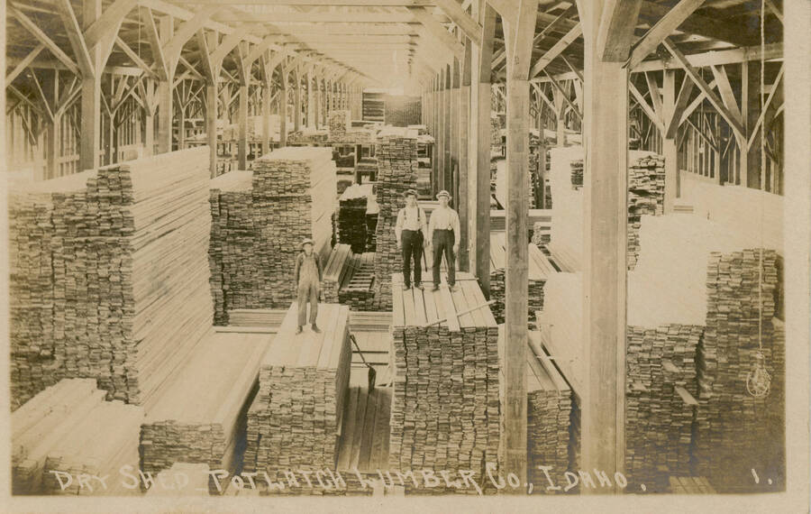 Postcard of the interior of the Dry Shed at the Potlatch Mill with men standing on lumber piles.