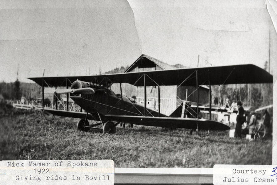 Nick Mamer giving airplane rides in Bovill. Many people can be seen standing around the airplane, which is parked in front of a building.