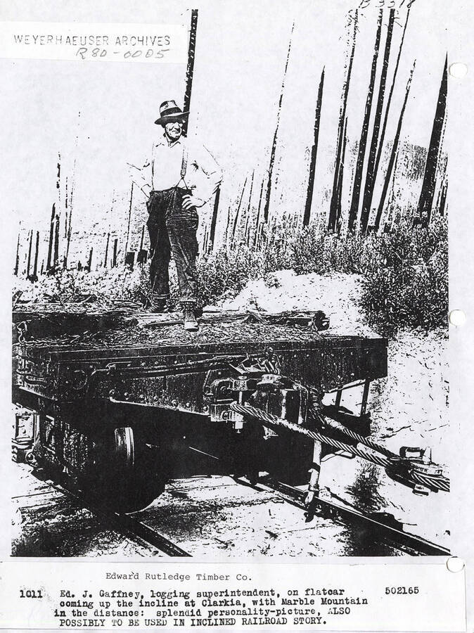 Edward J. Gaffney, the logging superintendent, standing on a flatcar. The photo was taken as the flatcar was coming to an incline in Clarkia. Marble Mountain can also be seen in the distance.