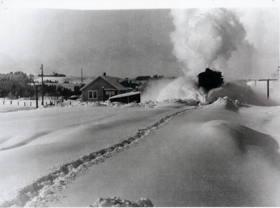 Photograph of the WI&M Railway steam locomotive, pulling empty log cars, plowing through snow at Princeton on its way to Bovill.