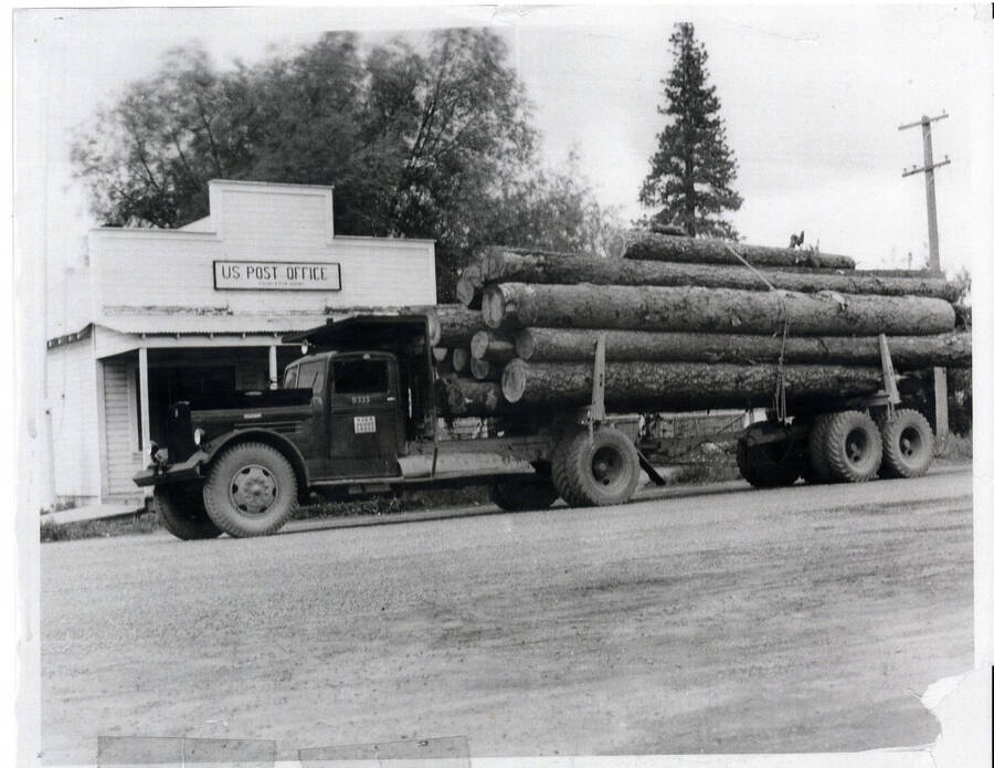 Photograph of logging truck on the way to the Potlatch Mill in front of the Princeton Post Office.