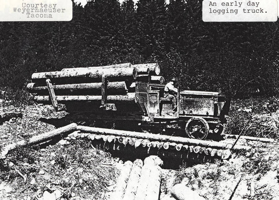 An early day logging truck. The truck has a stack of logs piled in the back and a man driving in the front.