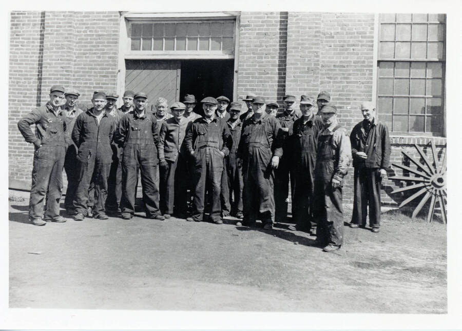 Photograph of (left to right) Kenneth King, Alfred Howard, Herman Bergman, Henry Young, Lawrence Rogers, Ivan Joe Asterlund, Phil Tice, William (Bill) Anderson, Russel Young, Ole Lillyman, Shelton Andrew, Ernie Cameron, George Morsching, Roy Sorbie, Ernie Donaman, Bill Schmitt, Conrad "Ole" Osterlund, Wesley Hoover, Bill Howard, Billie Doyle standing in front of the machine shop at the Potlatch Mill.