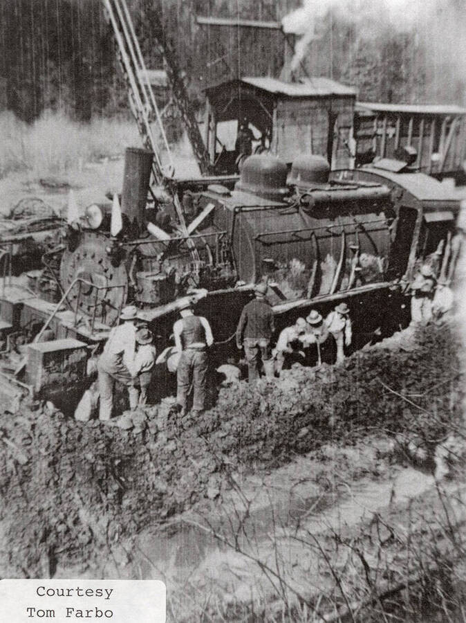 A group of men standing next to the side of a locomotive. A steam shovel can be seen behind the locomotive.
