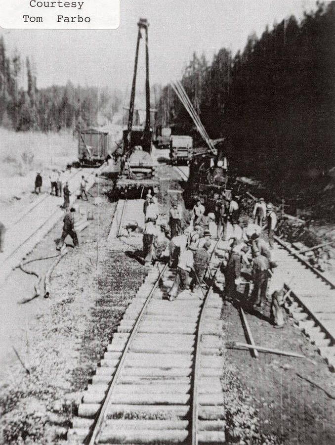 A group of men working on constructing the railroad. A locomotive, railroad cars, and a steam shovel can be seen sitting on the tracks behind the men.