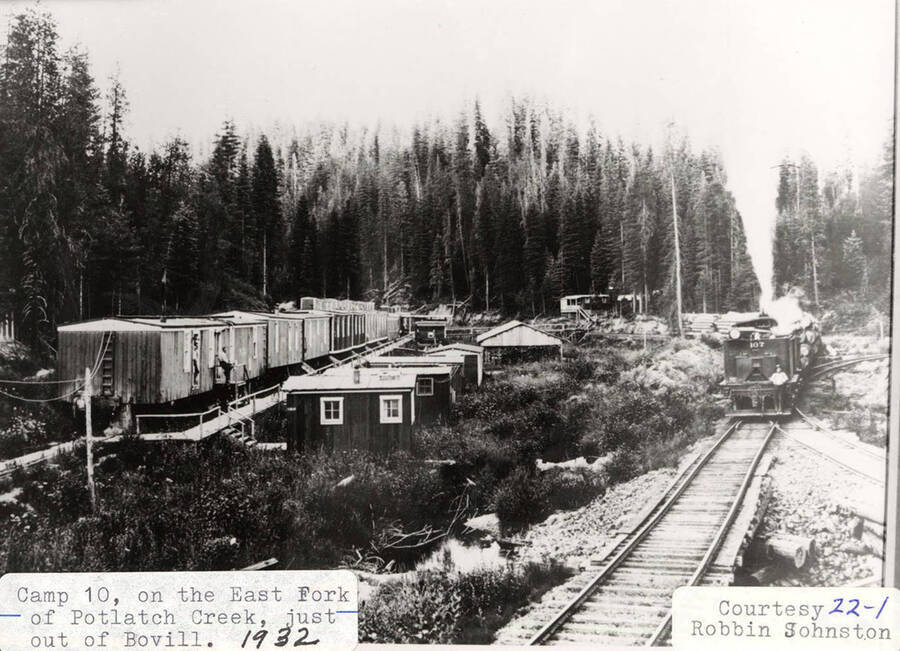 View of Camp 10, which is located on the East Fork of Potlatch Creek, just outside of Bovill. A No. 107 locomotive can be seen on the railroad tracks to the right of the camp's buildings.