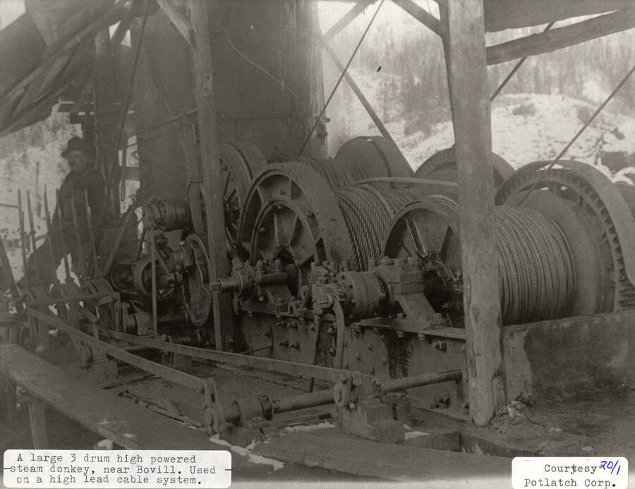 View of a large 3 drum high powered steam donkey, located near Bovill, Idaho. The machine was used on a high lead cable system. A man can be seen leaning up against the equipment.
