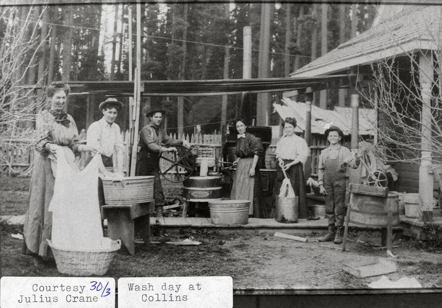Three women, two men, and a boy can be seen washing laundry during wash day at the Collins area. They can be seen using washtubs and a washboard.