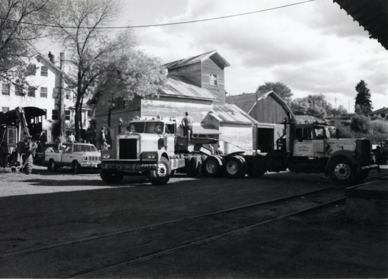 Photograph of trucks preparing to move Locomotive #1 to Scenic Six Park with City Hall, Creamery, and Produce Shed in the background.