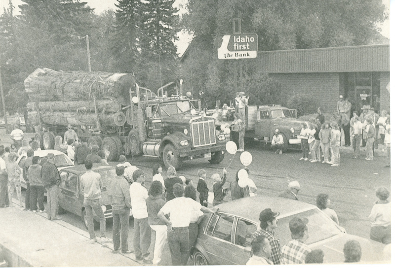 Photograph of people watching a logging truck in the Potlatch Days parade with bank building in the background.