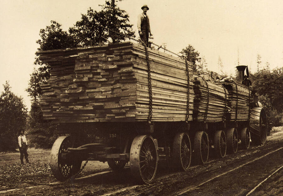 A tractor with stacks of logs chained to flat cars. A man can be seen standing on top of the stacks of logs and another man can be seen standing next to the cars.
