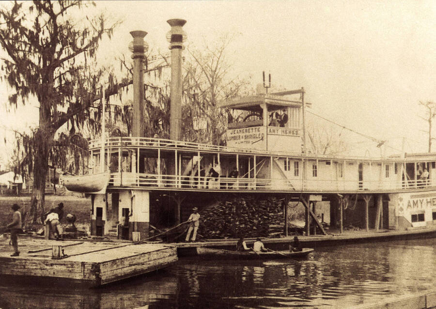 View of a steamboat being loaded with stacks of logs. A few men can be seen sitting in a canoe next to the boat.