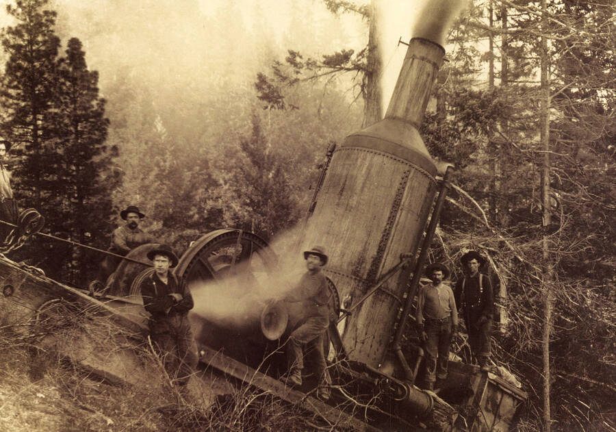 Men standing around a piece of logging equipment. Steam and can be seen coming out of the pipe at the top.