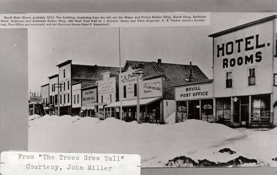 View of Main Street in Bovill during winter. Many shops can be seen on the street. Beginning from the left, the shops are Major and Peters Barber Shop, Bovill Drug, Spokane Hotel, Anderson and Anderson Barber Shop, Idle Hour Pool Hall, Grant and Giles Dry goods, E. K. Parker Menu's Furnishings, Post Office and Newsstand, and Sherman House.