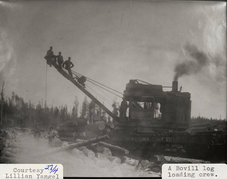View of a Bovill log loading crew. Men can be seen sitting on the equipment and standing next to a stack of logs.