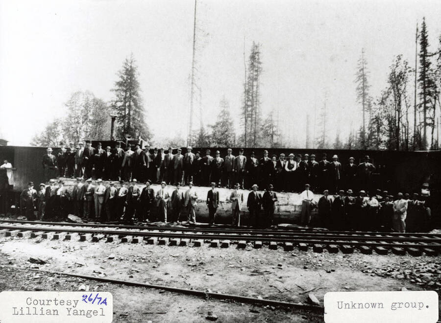 Photo of a group of men. The men are all wearing hats and are standing behind railroad tracks.