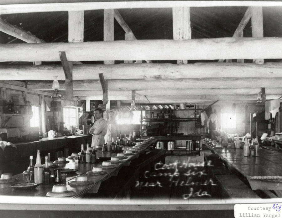 View of the cookhouse at Camp 6. Two men in aprons can be seen standing next to tables that have dishes stacked on them.
