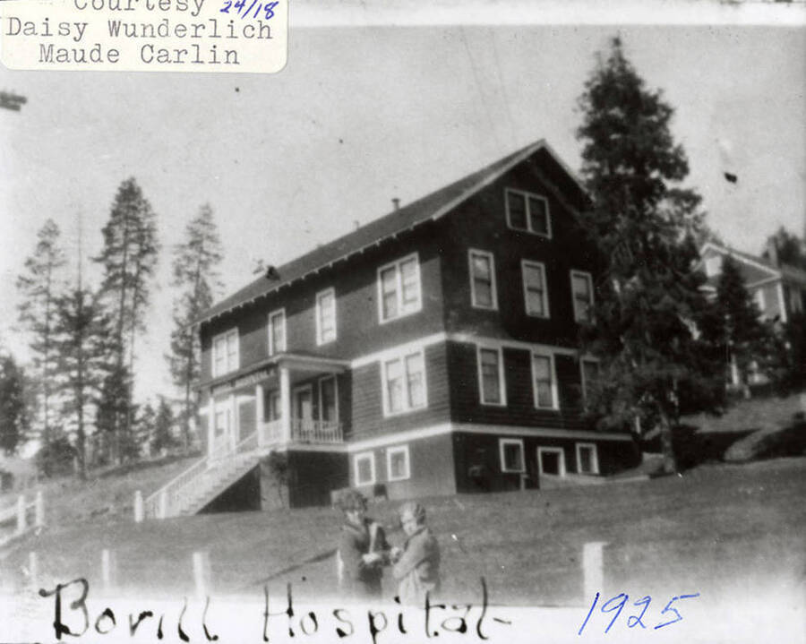 View of the hospital in Bovill, ID. Two people can be seen standing on the street outside the hospital and a building can be seen behind it.