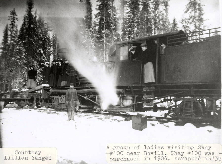 A group of ladies visiting the Shay No. 100 locomotive near Bovill, Idaho. The locomotive was purchased in 1906 and scrapped in 1939. A group of ladies can be seen standing on the locomotive and man can be seen standing next to it in the snow.