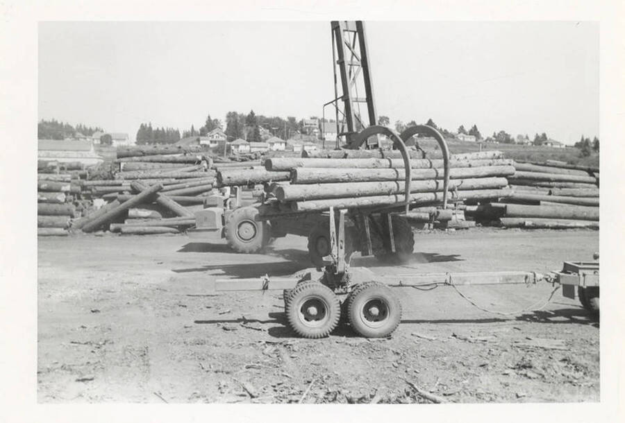 A front view of the LeTourneau log unloading machine with grappling- like hooks in front of several stacks of logs and the town.