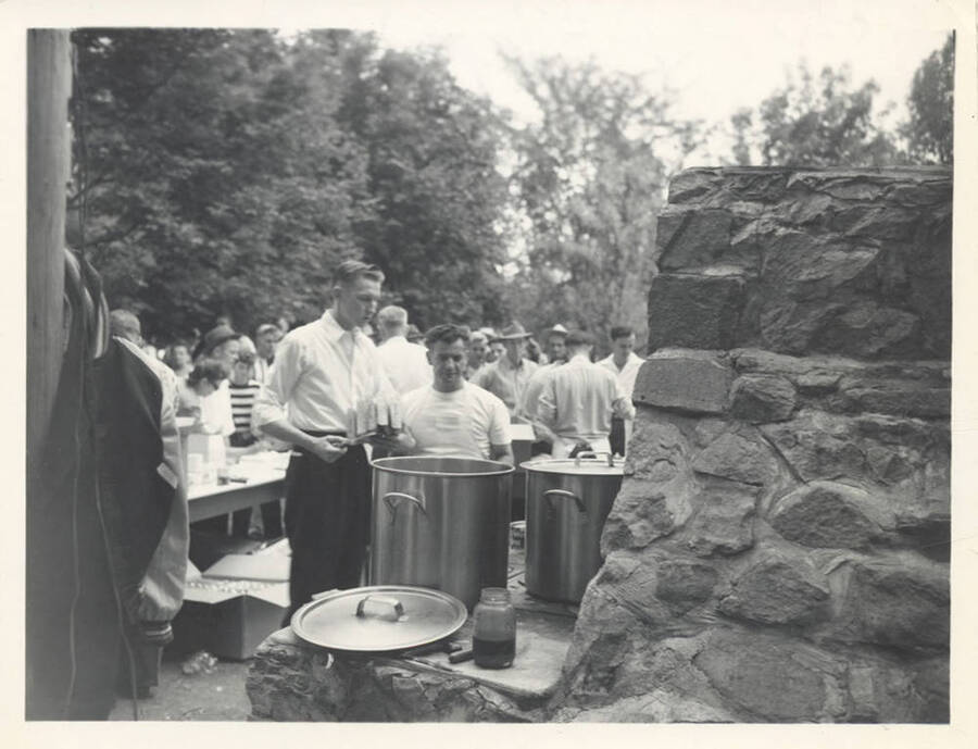 A view of the food cooking in pots while a large group of people wait eagerly behind.