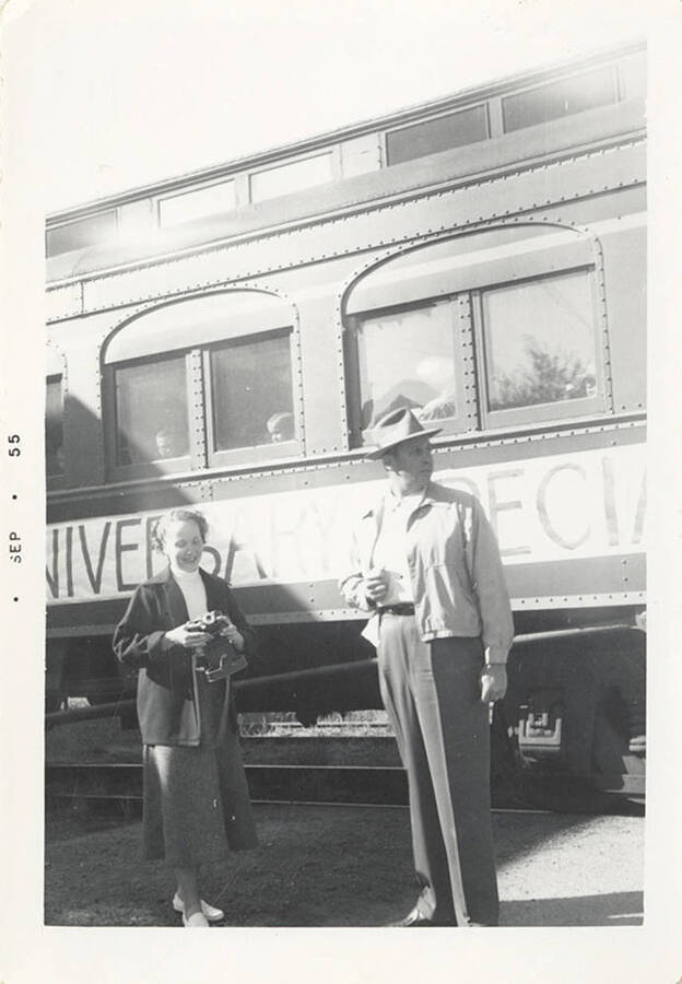 A man and a woman with a camera outside the passenger car with the banner for the Potlatch 50th Anniversary Special. You can see children's heads in the windows.