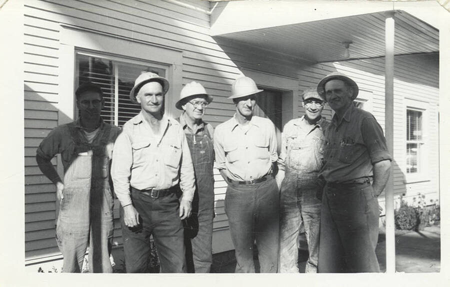 A group of six men wearing hats in front of a building.