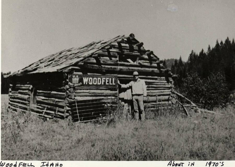 A man standing outside a hand built log building, the Woodfell, Idaho, United States Post Office. Photograph taken around 1970.