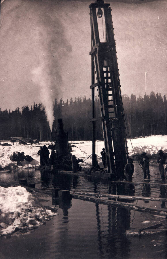 Employees in the log pond using a steam powered machine to lift logs out of the water.