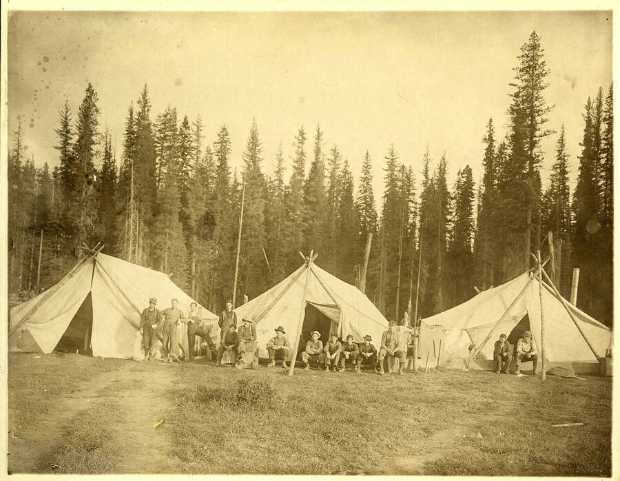 Men posing for the photo outside of three tents made from logs and canvas.